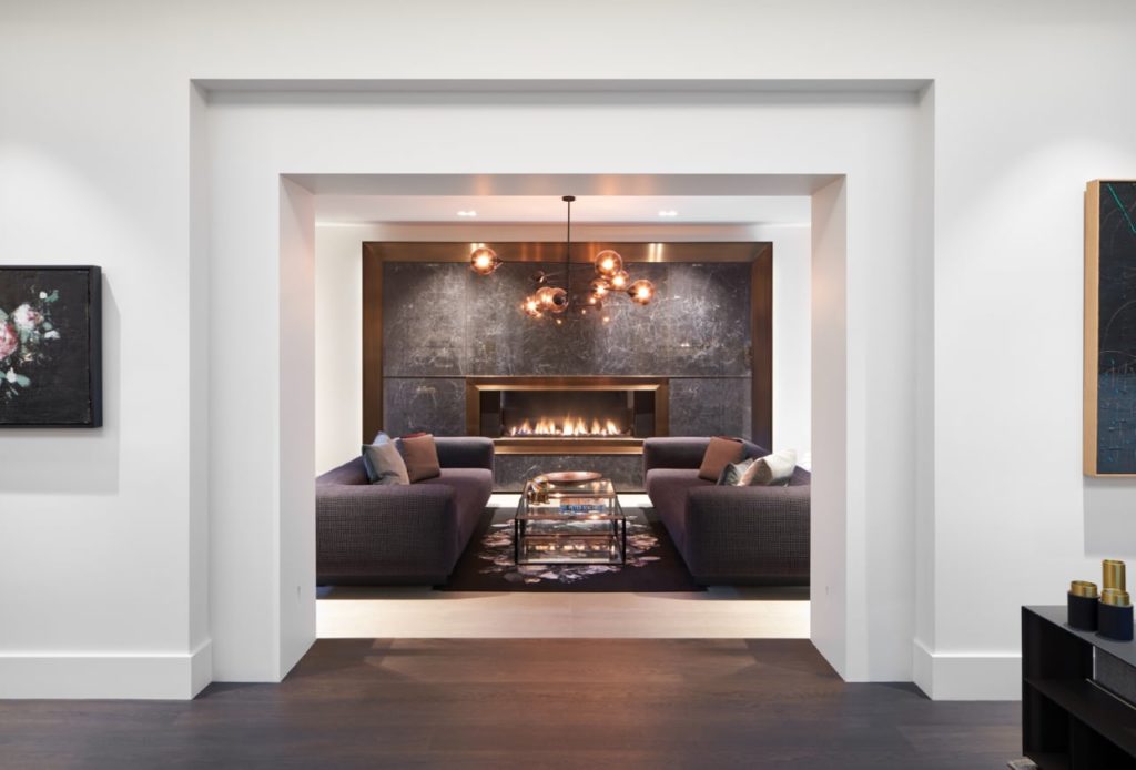 Linear gas fireplace is inset into a dramatic wall of dark veined stone trimmed in bronze.